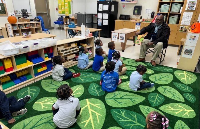  Dr. Kirkland Reading a story to students sitting around him on the carpet in the classroom.